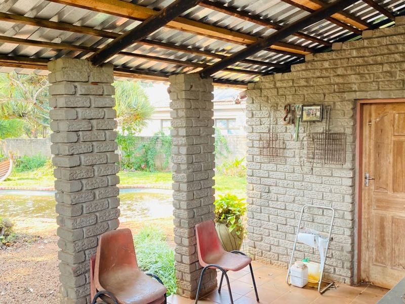 A covered outdoor patio area featuring exposed brick columns, a corrugated metal roof, and tiled flooring, overlooking the garden and pool area.