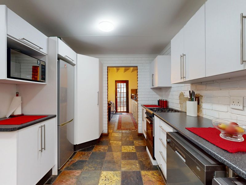 A functional kitchen featuring white cabinetry, dark laminate countertops, slate tile flooring, and exposed brick walls, with integrated appliances and direct access to an adjacent room.