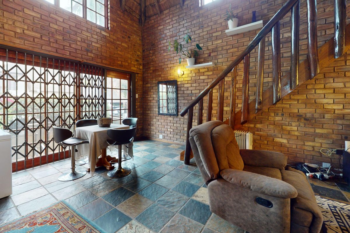 Spacious living area featuring exposed brick walls, slate tile flooring, and a timber staircase under a high thatch ceiling with sliding door access.