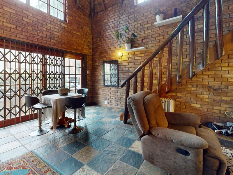 Spacious living area featuring exposed brick walls, slate tile flooring, and a timber staircase under a high thatch ceiling with sliding door access.