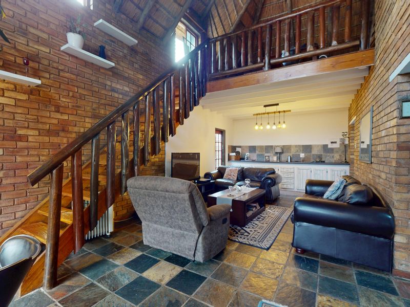 Open-plan living area featuring exposed brick walls, a thatched roof, slate flooring, and a timber staircase leading to a mezzanine level.
