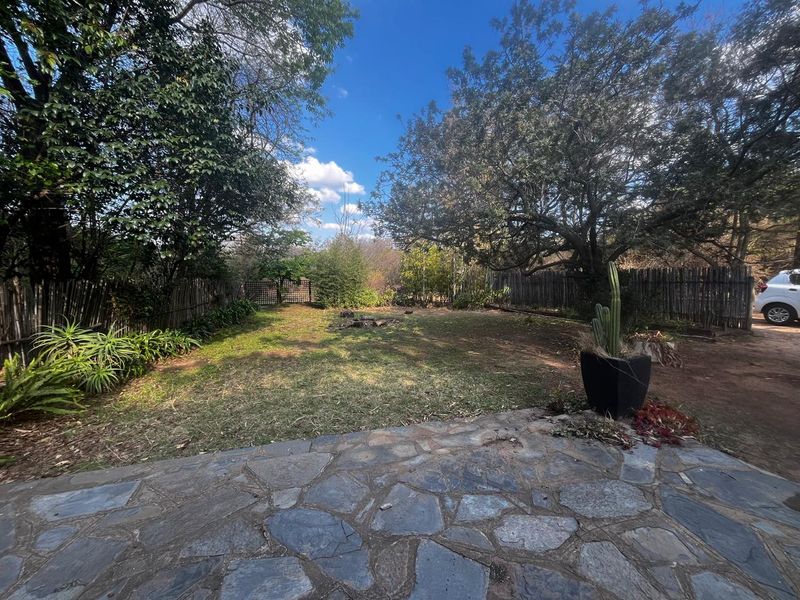 A spacious rear garden featuring a natural stone paved patio, mature trees, and rustic wooden pole fencing.