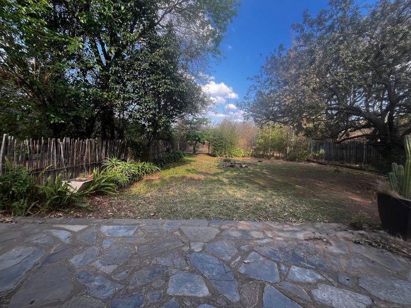 A spacious backyard garden featuring a natural stone paved patio, established trees, and perimeter wooden pole fencing.