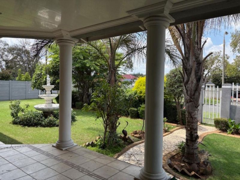 A covered patio featuring classical columns, overlooking a well-maintained garden with a stone walkway and a decorative fountain.