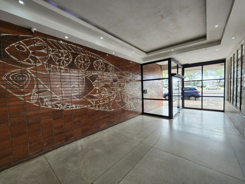 A spacious apartment building entrance lobby featuring a decorative brick wall, terrazzo flooring, and large glass doors leading to the street.