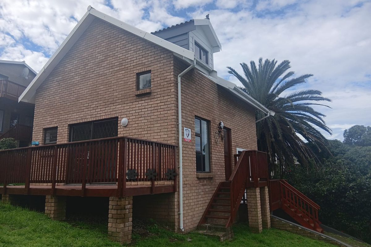 A rear view of a brick cottage featuring a raised timber deck, external staircase, and a dormer window set within a landscaped garden.