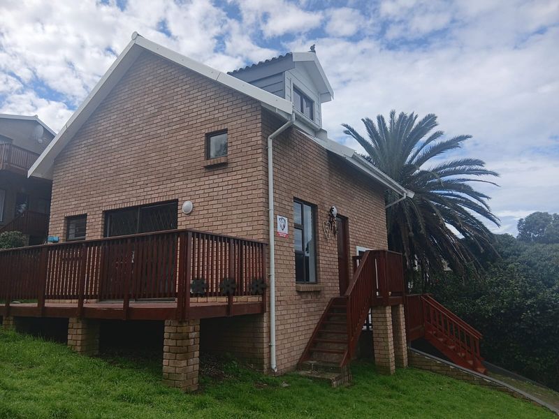 A rear view of a brick cottage featuring a raised timber deck, external staircase, and a dormer window set within a landscaped garden.