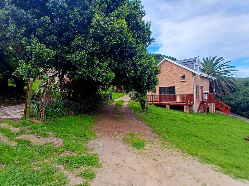 A rear view of a brick cottage featuring a raised timber deck, staircase, and surrounding garden area with mature trees.
