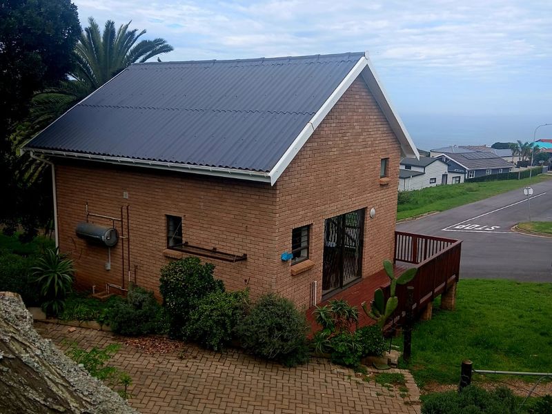 Exterior view of a brick cottage featuring a corrugated metal roof, a raised timber deck, and paved driveway access in a coastal setting.