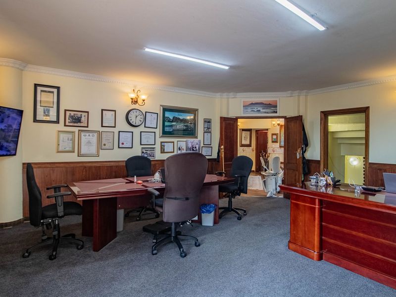 A spacious home office featuring timber wall paneling, decorative cornices, and multiple lighting fixtures, providing a functional workspace with access to adjacent rooms.