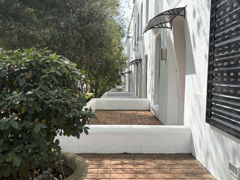 A perspective view of the townhouse exterior facade featuring brick paved walkways, low masonry walls, and protective awnings over the entrances.