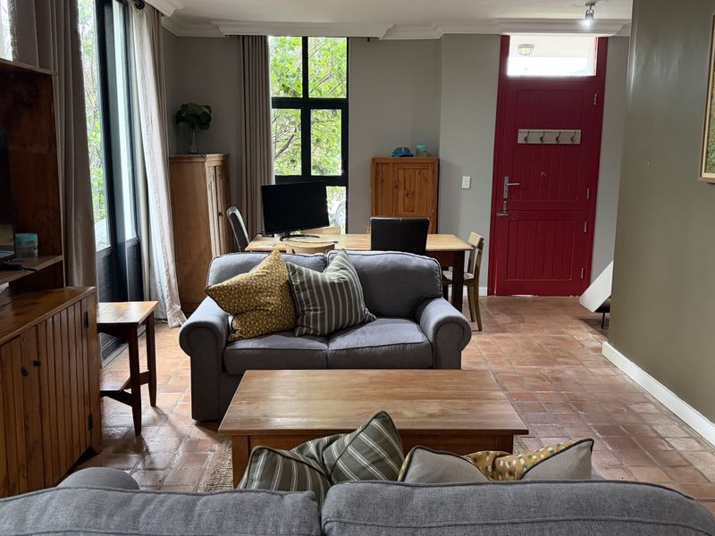 A living room featuring terracotta floor tiles, a large window, and a distinctive red entrance door, offering a warm and inviting atmosphere.