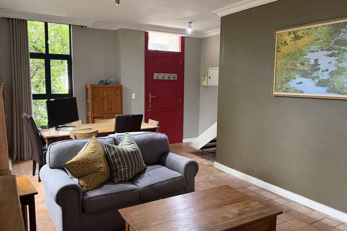 A spacious living area featuring terracotta tile flooring, large windows providing natural light, and a distinctive red entrance door with classic cornice detailing.