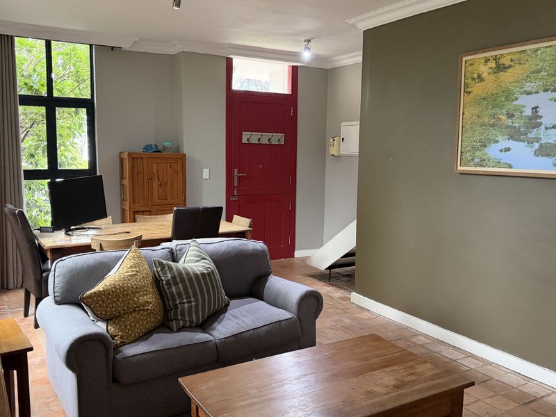 A spacious living area featuring terracotta tile flooring, large windows providing natural light, and a distinctive red entrance door with classic cornice detailing.