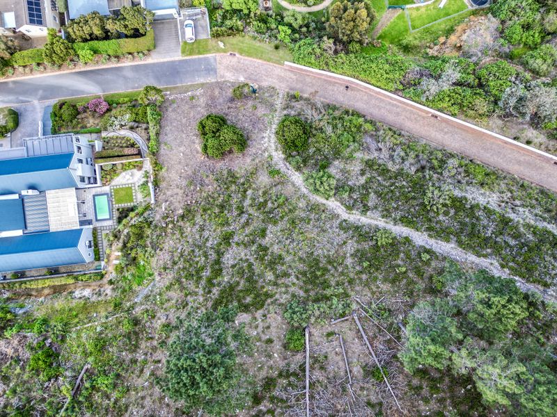 Aerial view of a vacant residential plot featuring natural vegetation, sloped terrain, and adjacent paved road access.