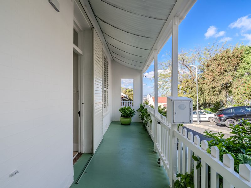 A covered front veranda featuring a painted concrete floor, white picket fencing, and a corrugated metal roof, providing a charming street-facing entrance area.
