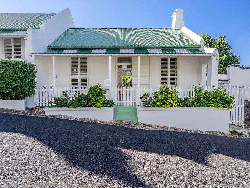 Charming cottage-style home featuring a classic white facade, green corrugated roof, covered veranda, and a neat picket fence with landscaped garden beds.