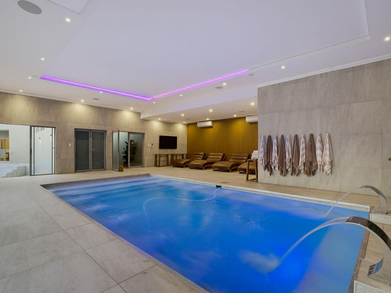A luxurious indoor pool area featuring modern tiled finishes, integrated water features, ambient LED lighting, and direct access through sliding glass doors.
