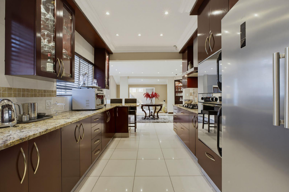 Modern galley-style kitchen featuring dark wood cabinetry, granite countertops, high-gloss tiled floors, and integrated appliances, flowing into an open-plan living area.