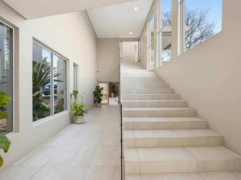 A bright and spacious modern hallway featuring a tiled staircase with a glass balustrade, large windows, and neutral finishes.