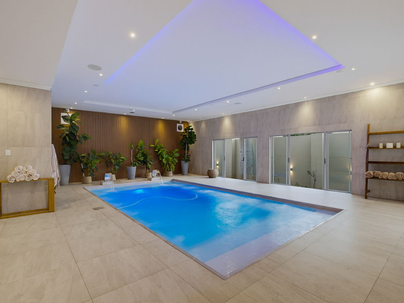 Luxurious indoor swimming pool area featuring modern tiled flooring, ambient LED cove lighting, and elegant wood slat wall paneling with direct access to outdoors.