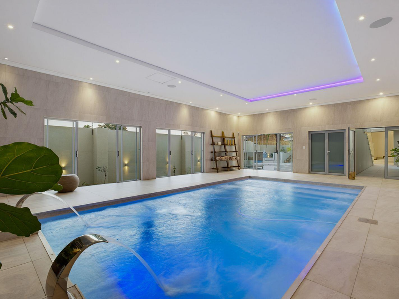 Luxurious indoor swimming pool area featuring modern tiled finishes, ambient LED ceiling lighting, and large sliding glass doors opening to the exterior.