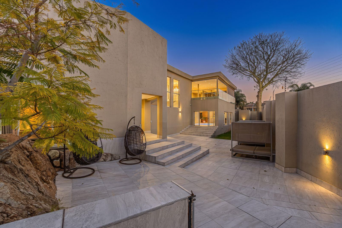 A modern rear exterior featuring a spacious tiled patio, stone retaining wall, and a glass-balustrade balcony overlooking the outdoor entertainment area.