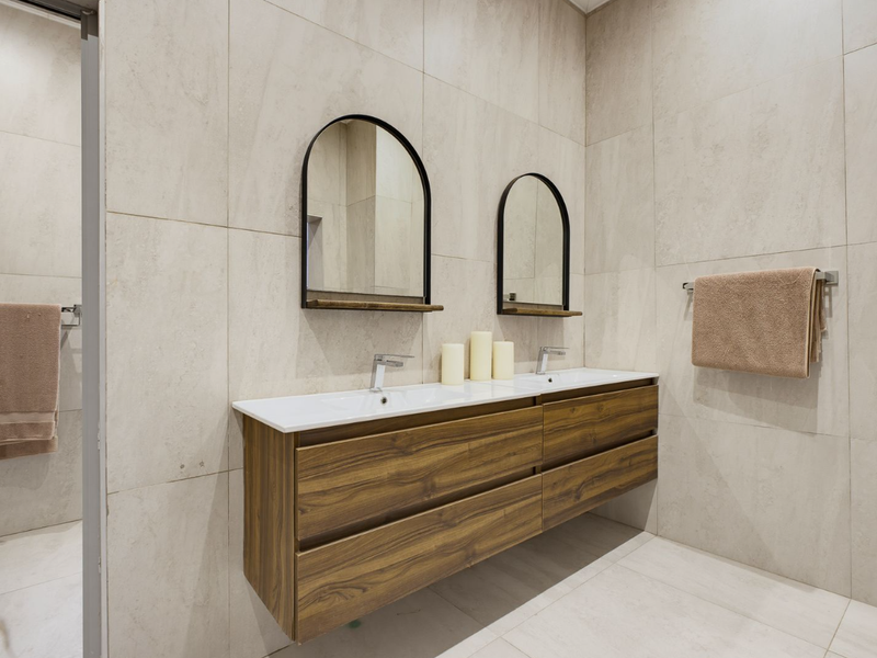 Modern bathroom featuring a sleek double vanity with wood-finish cabinetry, integrated sinks, chrome fixtures, and elegant arched mirrors against large-format neutral wall tiles.