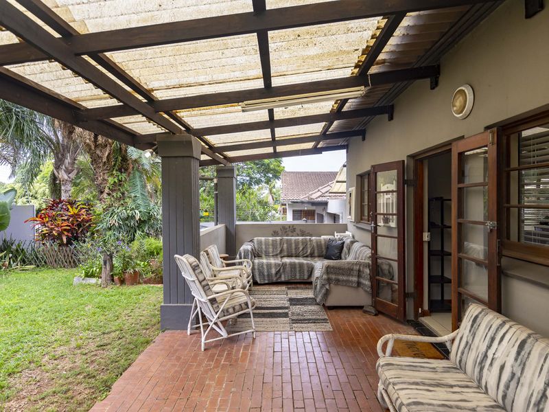 A covered outdoor patio featuring brick flooring, timber support beams, and translucent roof sheeting, providing direct access to the garden area.