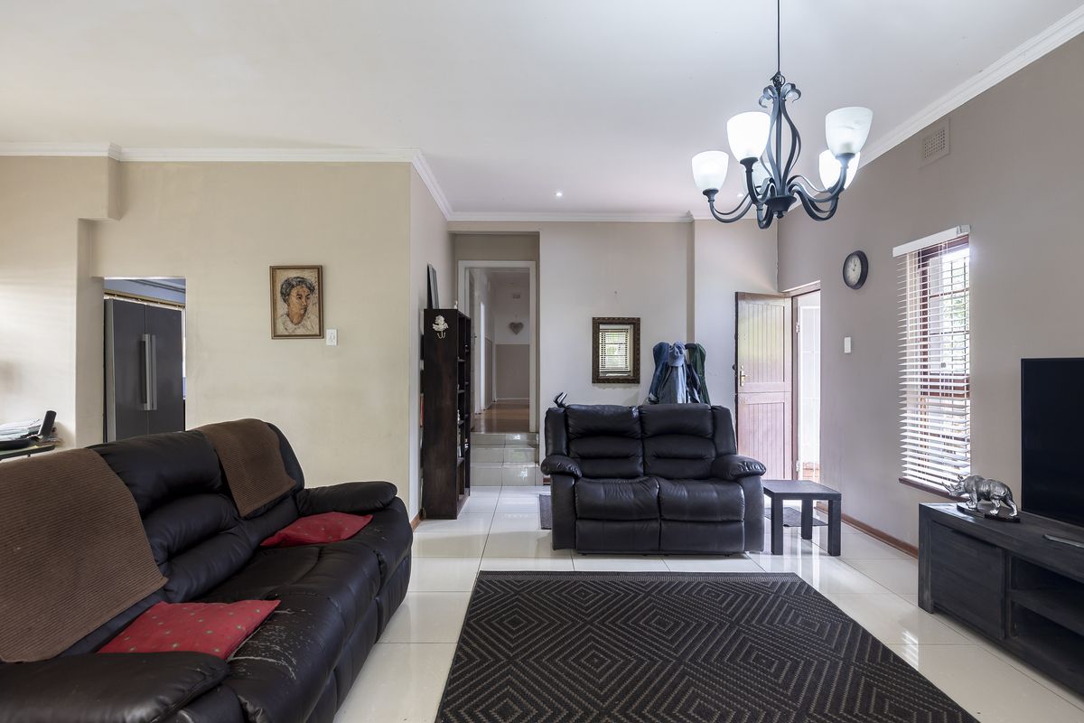 A spacious living room featuring tiled flooring, a decorative chandelier, and natural light through venetian blinds, leading to an internal hallway.
