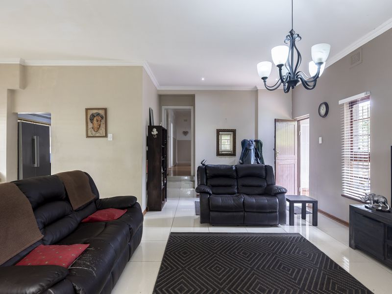 A spacious living room featuring tiled flooring, a decorative chandelier, and natural light through venetian blinds, leading to an internal hallway.