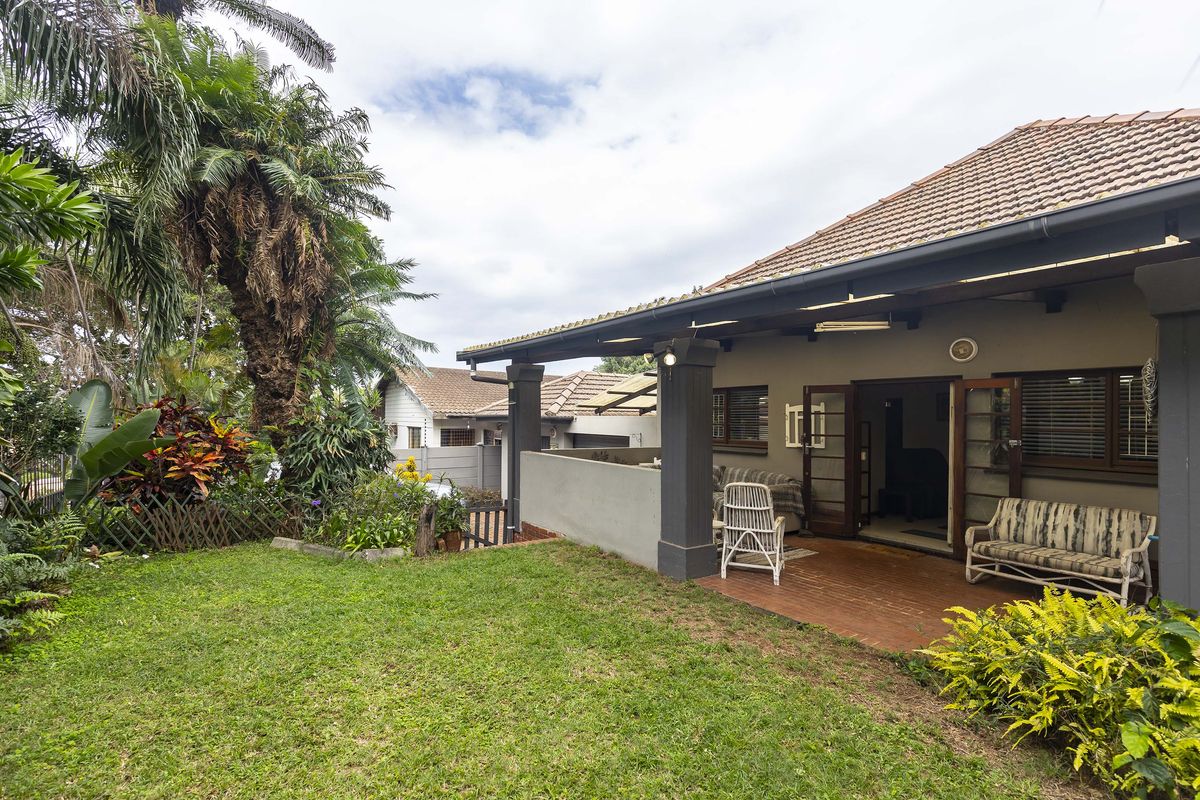 A well-maintained rear exterior view featuring a covered patio with brick flooring, concrete pillars, and a lush landscaped garden.