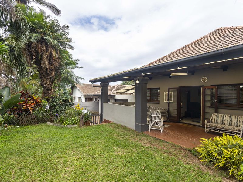 A well-maintained rear exterior view featuring a covered patio with brick flooring, concrete pillars, and a lush landscaped garden.