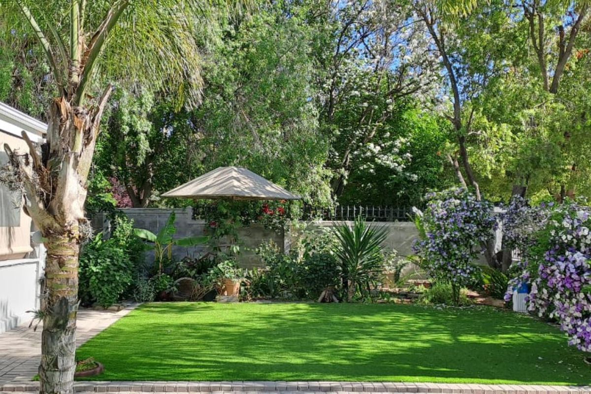 A well-maintained rear garden featuring a manicured lawn, established trees, flowering shrubs, and a paved patio area adjacent to the house.