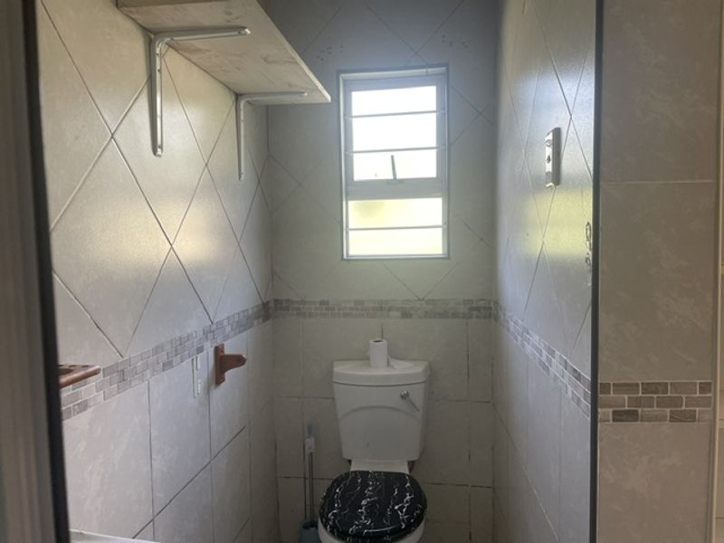 A compact bathroom featuring full-height wall tiling, a standard toilet, and wall-mounted shelving under a window with security bars.