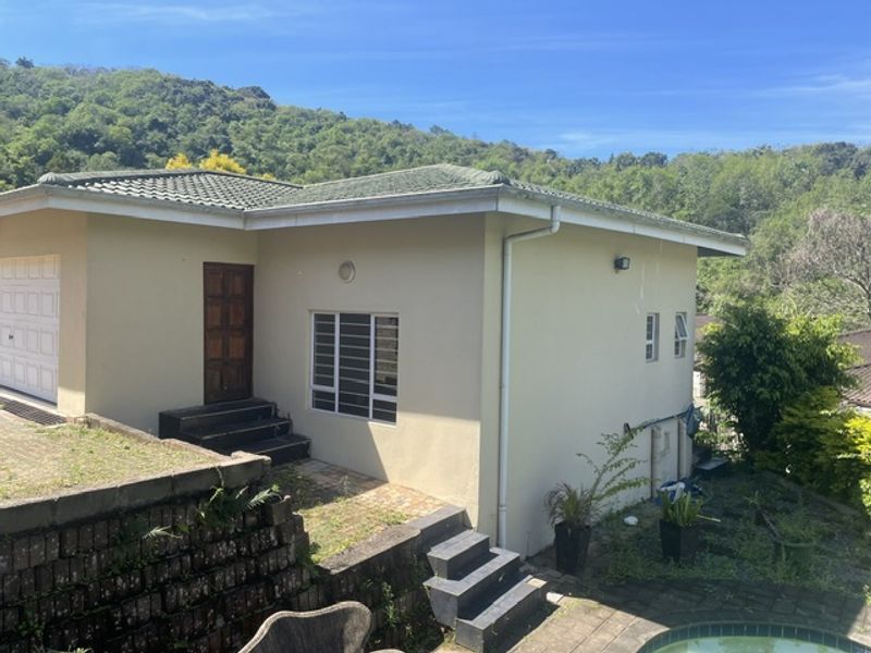 Rear exterior view of the cottage featuring a paved patio area, swimming pool, and tiered retaining walls set against a hillside backdrop.