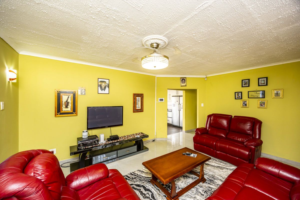 A living room featuring tiled flooring, a ceiling fan with a decorative rose, and wall sconce lighting, with a view into the adjacent kitchen area.