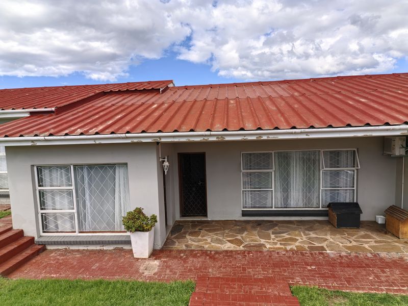 Front elevation of a single-story house featuring a red corrugated metal roof, stone-paved patio, and brick-paved walkway with security-fitted windows.