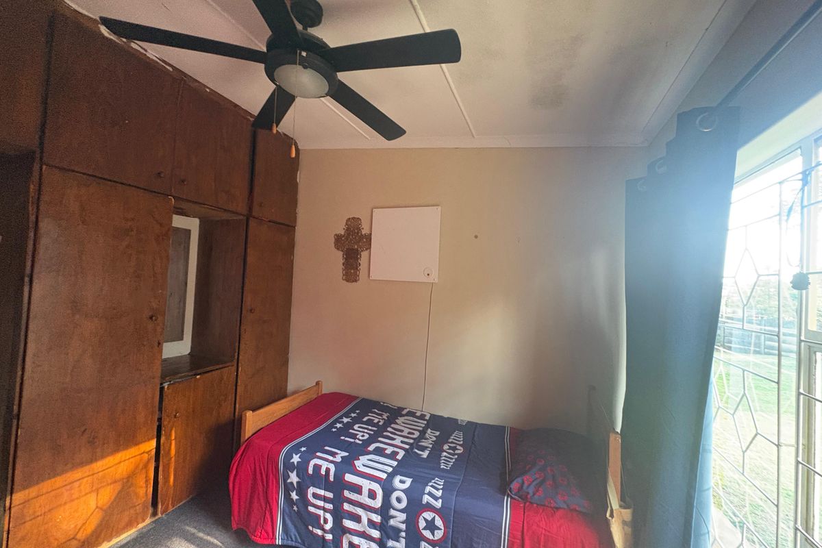 A bedroom featuring built-in wooden cupboards, a ceiling fan, and a window with security bars, showing signs of ceiling water damage.