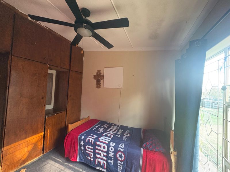 A bedroom featuring built-in wooden cupboards, a ceiling fan, and a window with security bars, showing signs of ceiling water damage.