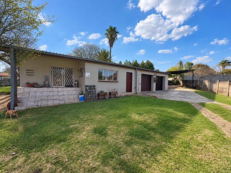 Rear view of the property featuring a tiled patio, double garage, paved driveway, and a spacious lawn enclosed by a brick boundary wall.