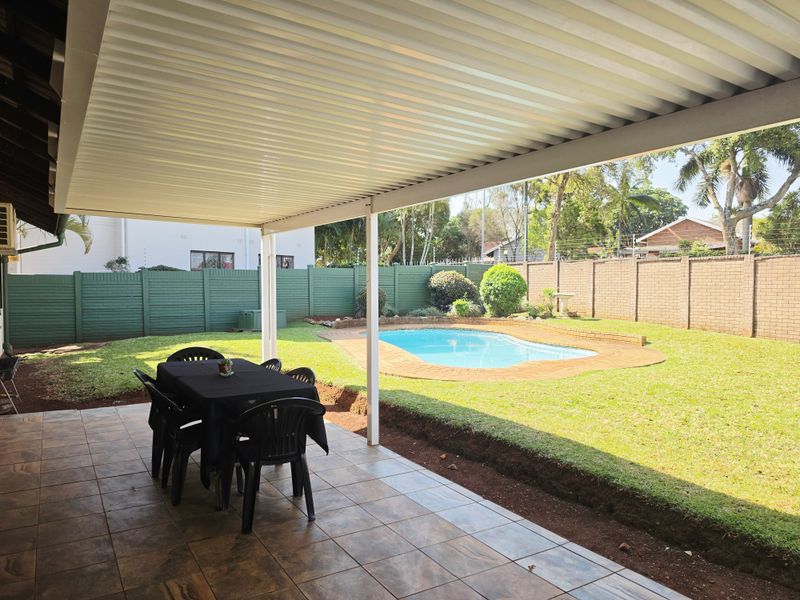 Covered patio area overlooking a private swimming pool and garden, featuring tiled flooring and a corrugated metal roof structure.