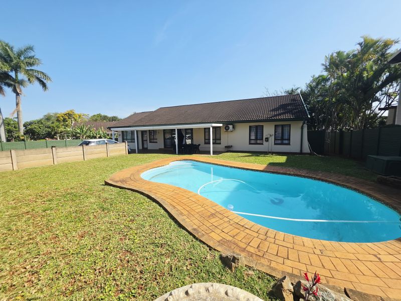 Rear view of the property featuring a swimming pool with brick coping, a covered patio area, and a well-maintained garden with boundary walling.
