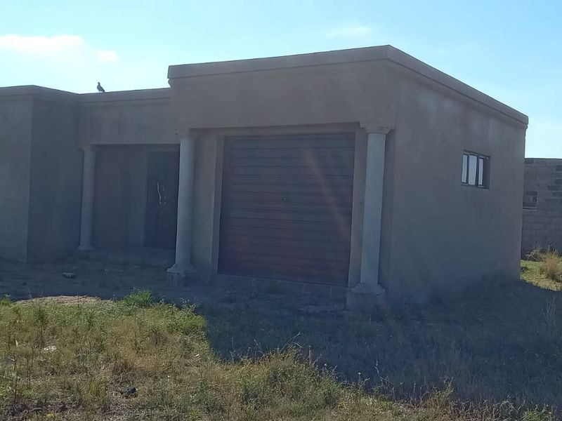 A single-story residential property under construction, featuring an integrated garage, unfinished exterior plaster, and surrounding undeveloped land.