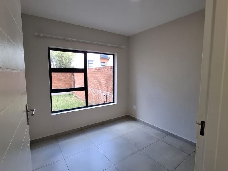 A bright, empty bedroom featuring neutral tiled flooring and a large aluminium-framed window overlooking the rear garden area.