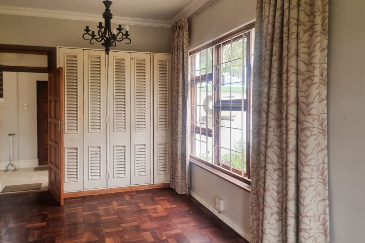 A spacious bedroom featuring classic parquet flooring, built-in louvered cupboards, and large windows with security bars, offering a bright and functional living space.