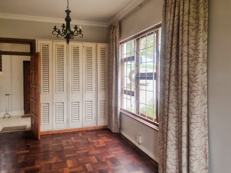 A spacious bedroom featuring classic parquet flooring, built-in louvered cupboards, and large windows with security bars, offering a bright and functional living space.