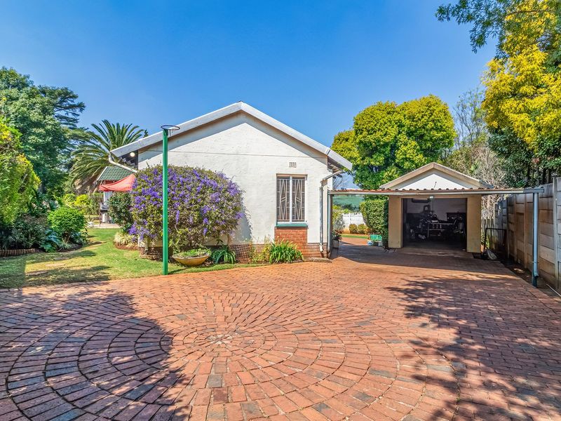 Front view of the property featuring a circular brick paved driveway, a single garage with an attached carport, and established garden surroundings.