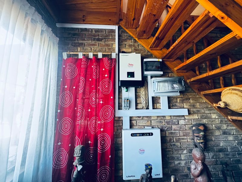 Utility area featuring a wall-mounted solar inverter, lithium battery system, and electrical distribution board installed against an exposed brick wall beneath a timber staircase.