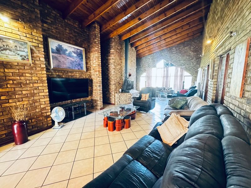 Spacious living room featuring high exposed timber beam ceilings, rustic facebrick walls, tiled flooring, and a prominent freestanding fireplace with a large arched window.